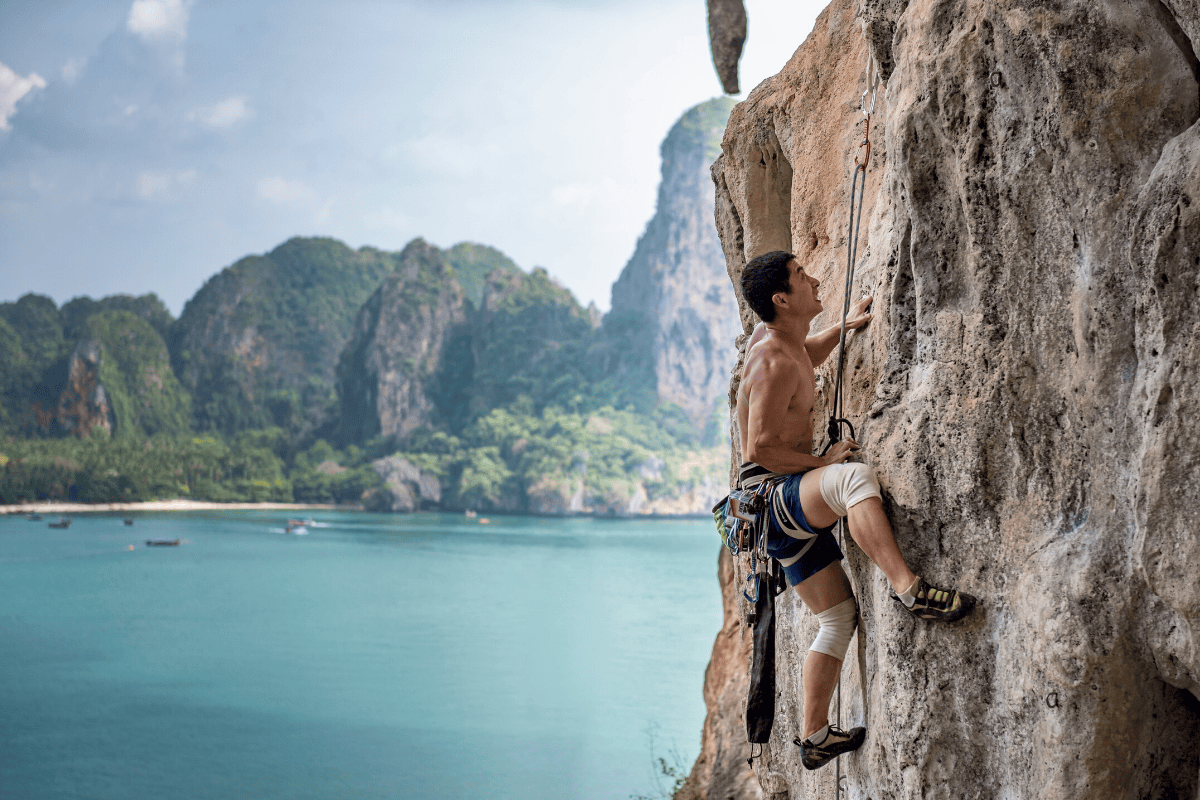 un homme escaladant une paroi rocheuse plate à l'aide de cordes de guidage, avec la plage de Railay et ses falaises de calcaire en arrière-plan à gauche