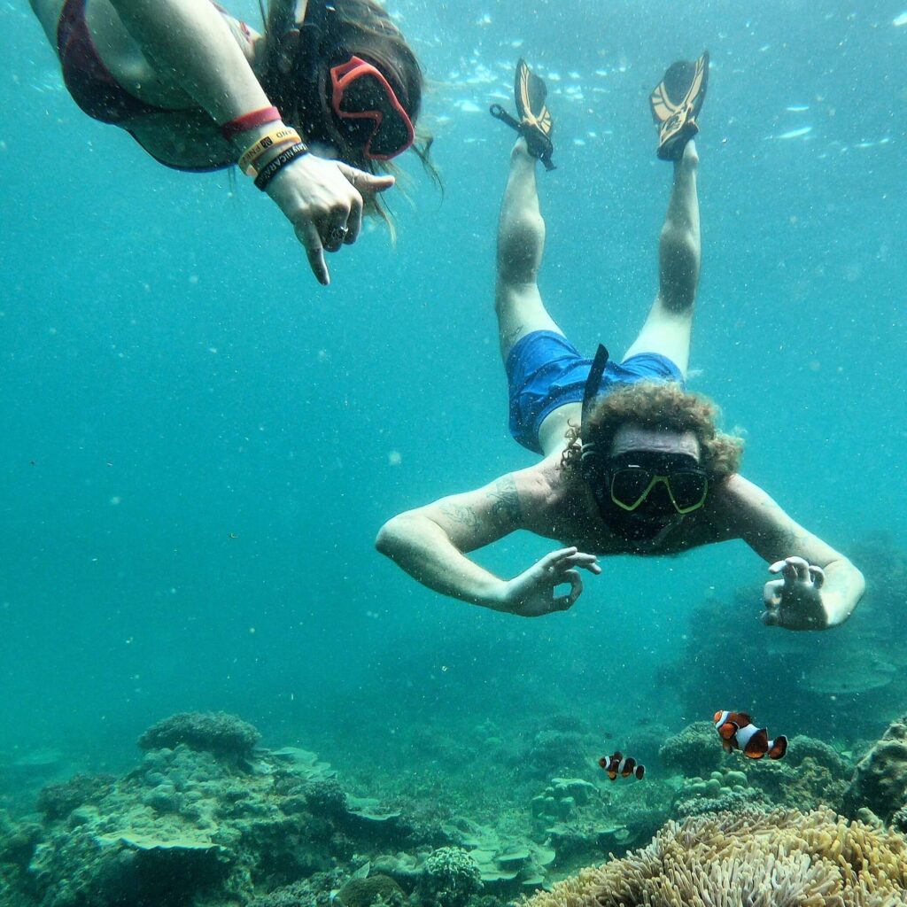 dos buceadores de snorkel con los pulgares hacia la cámara mientras nadan junto a un pez nemo (pez payaso)