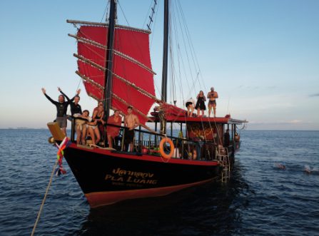 Guests posing and celebrating on the bow and roof of a red-sailed Thai junk boat during a sunset cruise in Krabi