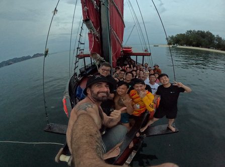 Private group selfie of smiling guests on the bow of a red-sailed junk boat during a Krabi sunset cruise