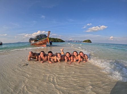 Guests lying in shallow turquoise water on a sandbar with a traditional longtail boat in the background