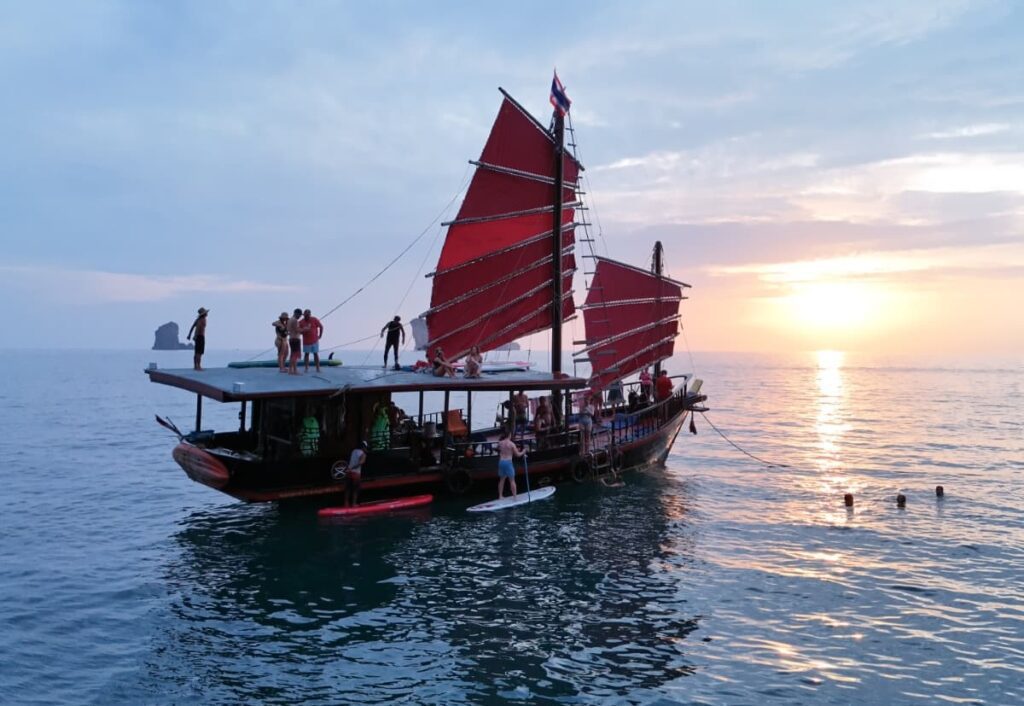 Crucero al atardecer en Krabi: un barco al atardecer con huéspedes practicando paddle surf junto a él y otros disfrutando de la puesta de sol desde la azotea