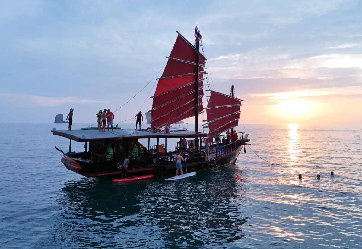 Crucero al atardecer en Krabi: un barco al atardecer con huéspedes practicando paddle surf junto a él y otros disfrutando de la puesta de sol desde la azotea