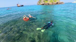 Un guía que lleva a dos personas por los arrecifes de Koh Ya Wa Sam en una boya salvavidas. Al fondo, el barco tradicional de Krabi Sunset Cruises.
