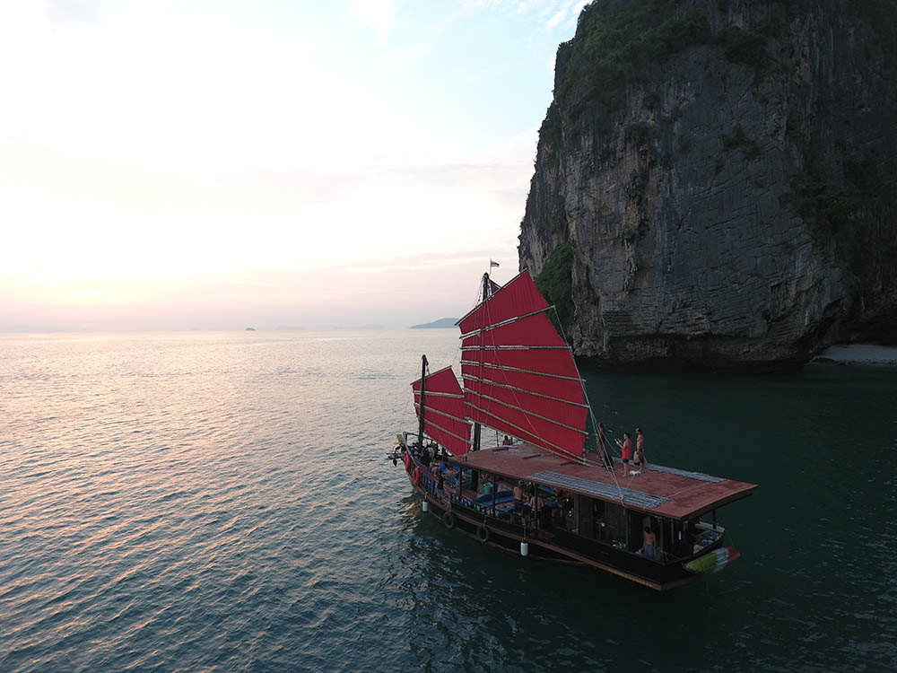 barco turístico de madera de 2 plantas en krabi navegando junto a un acantilado con sus velas rojas izadas al atardecer