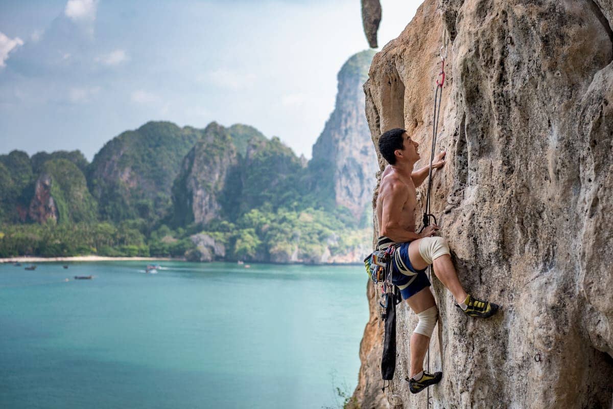 un hombre escalando una pared rocosa plana con cuerdas de guía, con la playa de Railay y sus acantilados de piedra caliza al fondo a la izquierda