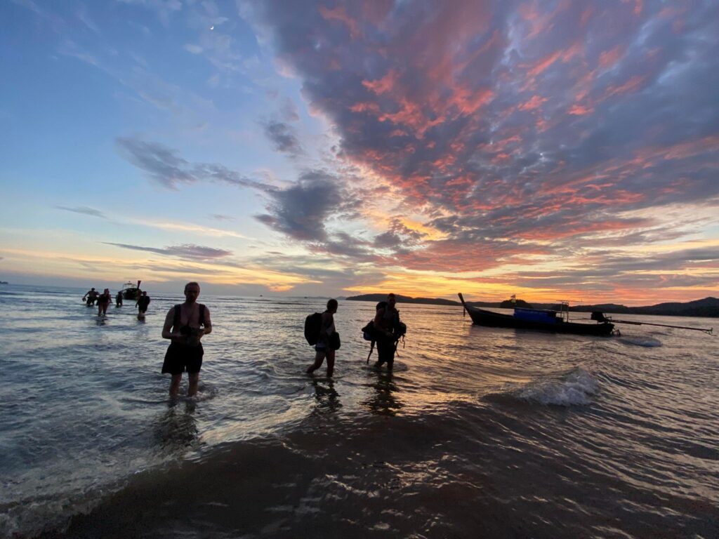 gente vadeando el agua durante la puesta de sol en la playa de aonang con nubes de colores morado y naranja