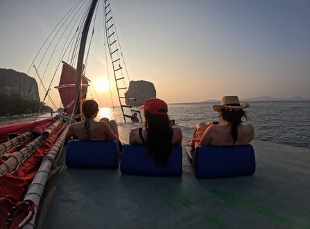 Tres mujeres descansan en la cubierta del barco, contemplando la puesta de sol tras los acantilados de piedra caliza