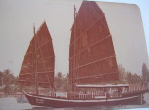 Ancienne photo du Pla Luang avec ses deux voiles rouges, amarré près de palmiers.